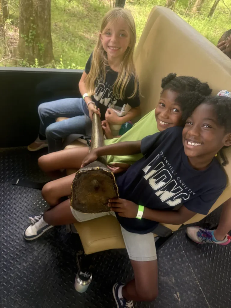 Three smiling young girls sitting on a tan bench seat in an open-sided vehicle, holding a large, brown, bone-like prop across their laps.