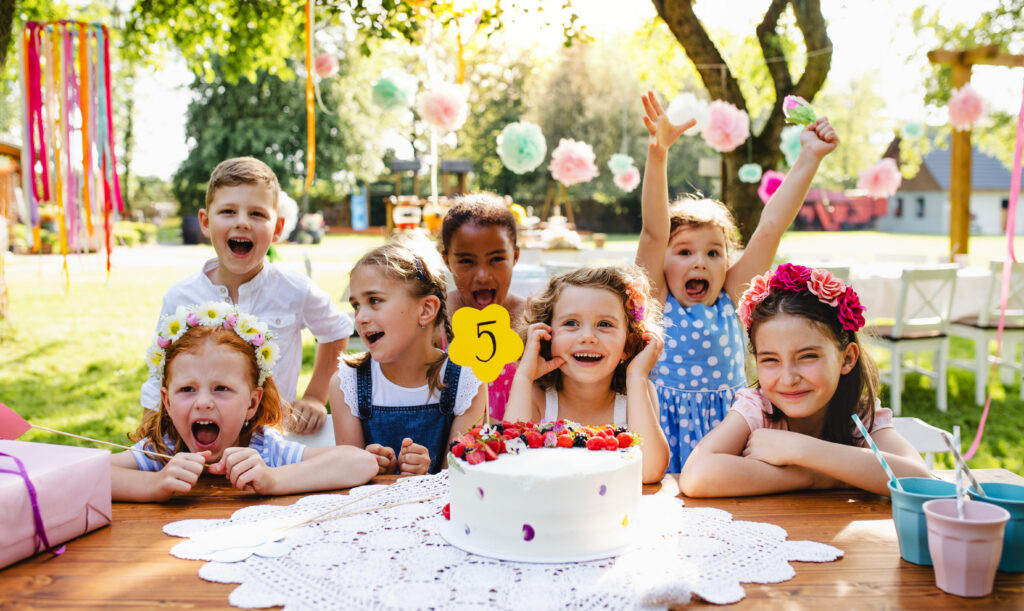 Children with cake standing around table on birthday party in garden in summer.